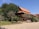 A traditional wooden building with a red-tiled roof, surrounded by lush greenery and trees. The architecture features open spaces and ornate design elements typical of Southeast Asian style. The area is peaceful, with a paved path in the foreground leading up to the structure.
