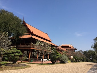 A traditional Asian architectural structure with ornate, sloping roofs is surrounded by a lush garden with neatly trimmed bushes and trees. The bright blue sky provides a clear backdrop, and the structure appears to be made of wood, giving a warm and welcoming vibe. A paved pathway leads up to the building, emphasizing its grandeur and accessibility.