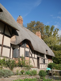 A traditional cottage featuring a thatched roof with wooden beams and brick accents. The building is surrounded by lush greenery and a small garden. A clear blue sky and a few trees in the background complete the picturesque setting.