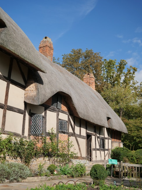A traditional cottage featuring a thatched roof with wooden beams and brick accents. The building is surrounded by lush greenery and a small garden. A clear blue sky and a few trees in the background complete the picturesque setting.