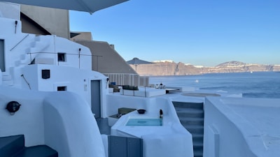 A serene coastal setting featuring whitewashed architecture characteristic of Mediterranean designs, with curved walls and stairways. A small pool or hot tub sits in the foreground, and the view extends to calm blue waters with distant cliffs under a clear blue sky. Minimal furniture and decor create a peaceful, relaxing atmosphere.
