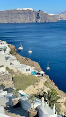 A coastal view of white buildings built into a rocky hillside overlooking a vivid blue sea. A few sailboats are anchored in the calm water, and in the background, a large cliff rises majestically. Lush greenery and palm trees are interspersed among the buildings, with a pool visible on one of the terraces.