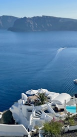A coastal scene featuring white, modern, cliffside architecture overlooking a serene blue sea. The structures include several terraces, a small swimming pool, and lush greenery, with a prominent umbrella-shaded seating area. In the distance, a boat leaves a wake as it moves across the calm water. The background has dark, rugged cliffs.
