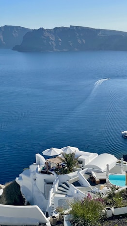 A coastal scene featuring white, modern, cliffside architecture overlooking a serene blue sea. The structures include several terraces, a small swimming pool, and lush greenery, with a prominent umbrella-shaded seating area. In the distance, a boat leaves a wake as it moves across the calm water. The background has dark, rugged cliffs.