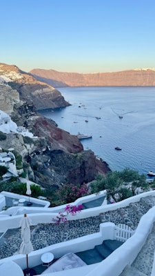 A picturesque coastal landscape featuring a steep cliffside dotted with white-washed buildings overlooking a tranquil blue sea. Several boats float peacefully in the water, surrounded by the warm colors of a sunset. The foreground includes terraced patios with outdoor seating, vibrant pink flowers, and green foliage.