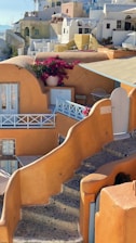 A picturesque scene featuring traditional Cycladic architecture with white and pastel-colored buildings, narrow stone stairways, and vibrant pink bougainvillea flowers in a pot. The buildings have curved roofs and small balconies, characteristic of a Mediterranean island setting.