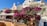 A picturesque scene featuring vibrant bougainvillea cascading down a hillside terrace among charming white and pastel-colored buildings. At the top, a Greek flag flutters in the breeze. Below, there is an outdoor seating area with tables and chairs under a white canopy.