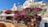 A picturesque scene featuring vibrant bougainvillea cascading down a hillside terrace among charming white and pastel-colored buildings. At the top, a Greek flag flutters in the breeze. Below, there is an outdoor seating area with tables and chairs under a white canopy.