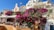 A picturesque scene featuring vibrant bougainvillea cascading down a hillside terrace among charming white and pastel-colored buildings. At the top, a Greek flag flutters in the breeze. Below, there is an outdoor seating area with tables and chairs under a white canopy.