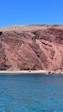 A vibrant coastline with a large, rugged red cliff rising above a sandy beach. The water in the foreground is a bright blue, contrasting with the earthy tones of the cliff. People are scattered along the beach, some under small umbrellas, enjoying the sunny day.