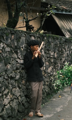 A person in traditional clothing stands against an old, stone wall playing a wooden flute. They are surrounded by greenery, with a few branches visible on the wall. The ground is paved, and the scene conveys a sense of calmness in a rural setting.