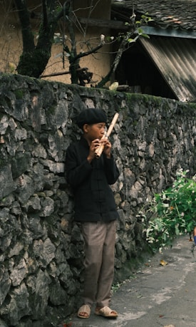 A person in traditional clothing stands against an old, stone wall playing a wooden flute. They are surrounded by greenery, with a few branches visible on the wall. The ground is paved, and the scene conveys a sense of calmness in a rural setting.