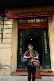 A person stands in front of a building entrance, with hands clasped together in a gesture resembling prayer or gratitude. They are carrying two backpacks and dressed casually. Above, a red banner with Vietnamese text spans the width of the entrance.