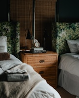 A cozy bedroom setting featuring two beds with white linens and patterned headboards. A wicker nightstand sits between the beds, adorned with a stack of books and a classic alarm clock. The room is softly lit, with two black bedside lamps flanking the nightstand. The neutral tones and plush textiles create a serene atmosphere.