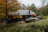 A cozy, secluded house nestled in a forested area with autumn foliage. The house is dark-colored with a sloped roof and features a spacious wooden deck with outdoor furniture. Warm interior lights are visible through the windows, suggesting an inviting atmosphere. The surrounding trees display vibrant shades of orange and green, with a manicured lawn in the foreground.