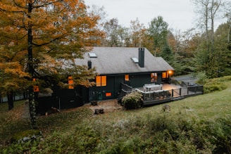 A cozy, secluded house nestled in a forested area with autumn foliage. The house is dark-colored with a sloped roof and features a spacious wooden deck with outdoor furniture. Warm interior lights are visible through the windows, suggesting an inviting atmosphere. The surrounding trees display vibrant shades of orange and green, with a manicured lawn in the foreground.
