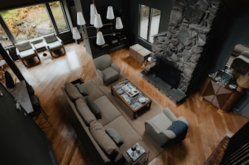 A cozy living room with a large sectional sofa and armchairs arranged around a stone fireplace. A coffee table with books and decor sits on a rug in the center. Large windows provide natural light, and pendant lights hang from the ceiling. Wooden floors and earth-tone colors create a warm atmosphere.