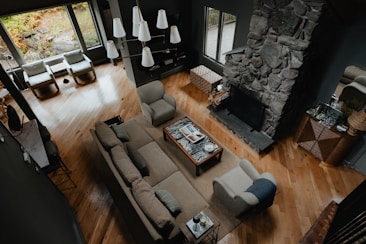 A cozy living room with a large sectional sofa and armchairs arranged around a stone fireplace. A coffee table with books and decor sits on a rug in the center. Large windows provide natural light, and pendant lights hang from the ceiling. Wooden floors and earth-tone colors create a warm atmosphere.