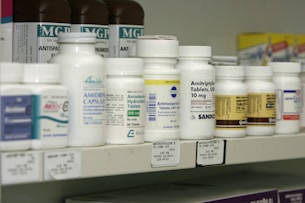A shelf filled with various bottles of medication, each labeled with different drug names and dosages. The labels are primarily white with some colorful accents. The bottles vary in size and shape, typical of a pharmacy or medical supply setting.