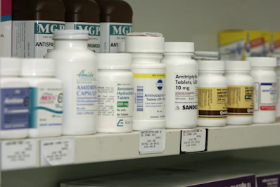 A shelf filled with various bottles of medication, each labeled with different drug names and dosages. The labels are primarily white with some colorful accents. The bottles vary in size and shape, typical of a pharmacy or medical supply setting.