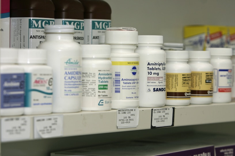 A shelf filled with various bottles of medication, each labeled with different drug names and dosages. The labels are primarily white with some colorful accents. The bottles vary in size and shape, typical of a pharmacy or medical supply setting.