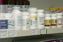 A shelf filled with various bottles of medication, each labeled with different drug names and dosages. The labels are primarily white with some colorful accents. The bottles vary in size and shape, typical of a pharmacy or medical supply setting.