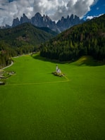 A small church is situated in the middle of a vast, vibrant green field, surrounded by dense forests and dramatic mountain peaks in the background. The sky is partly cloudy, adding depth and contrast to the scene.