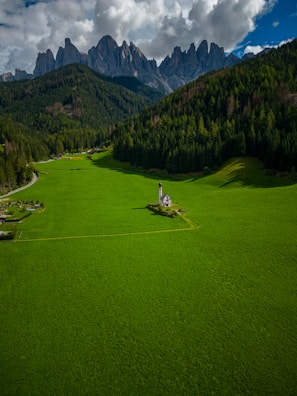A small church is situated in the middle of a vast, vibrant green field, surrounded by dense forests and dramatic mountain peaks in the background. The sky is partly cloudy, adding depth and contrast to the scene.