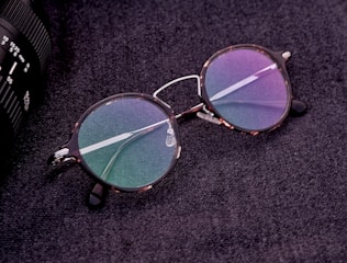 A pair of round eyeglasses with tortoiseshell frames and colorful reflective lenses is placed on a textured dark surface. The glasses are next to a part of a camera lens which is partially visible on the edge of the image.