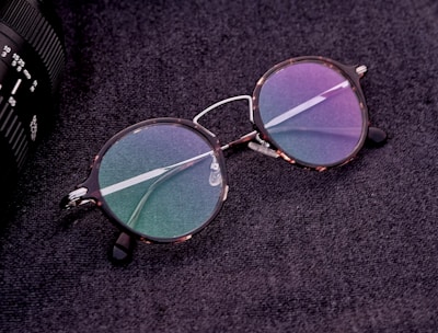 A pair of round eyeglasses with tortoiseshell frames and colorful reflective lenses is placed on a textured dark surface. The glasses are next to a part of a camera lens which is partially visible on the edge of the image.