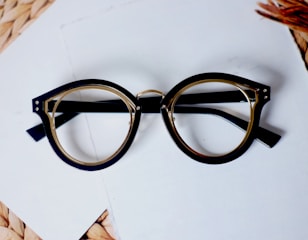 A pair of round, vintage-style eyeglasses with thick black frames rests on a white surface. There is a subtle gold detail around the lenses, and the wooden, woven texture of a mat is partially visible in the corner.