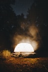 A glowing geodesic dome structure is situated in a dark forest clearing, emitting a warm light that contrasts with the surrounding trees and night sky.