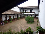 A traditional courtyard with a rustic, colonial-style building surrounding it. The structure has white walls and a clay tile roof with wooden beams supporting a covered walkway. There are potted plants and greenery along the periphery, and three people walking around. The ground is paved with hexagonal tiles, and there are balconies with potted plants on the second floor.