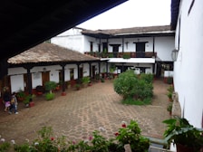 A traditional courtyard with a rustic, colonial-style building surrounding it. The structure has white walls and a clay tile roof with wooden beams supporting a covered walkway. There are potted plants and greenery along the periphery, and three people walking around. The ground is paved with hexagonal tiles, and there are balconies with potted plants on the second floor.