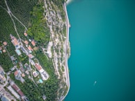 An aerial view of a coastal area featuring a winding road separating a densely forested hillside and a calm turquoise body of water. Residential buildings and green spaces are visible on the land side, with a clear divide between the urban area and the natural landscape.