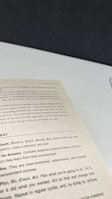 A close-up view of an open book page showing text on a desk or table. The text contains several sections with headers such as 'The Origins of Scrum' and various advice points, including observe, orient, decide, and act. The surface underneath the book is white with a dark background.