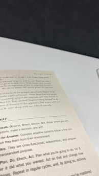 A close-up view of an open book page showing text on a desk or table. The text contains several sections with headers such as 'The Origins of Scrum' and various advice points, including observe, orient, decide, and act. The surface underneath the book is white with a dark background.