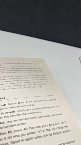 A close-up view of an open book page showing text on a desk or table. The text contains several sections with headers such as 'The Origins of Scrum' and various advice points, including observe, orient, decide, and act. The surface underneath the book is white with a dark background.