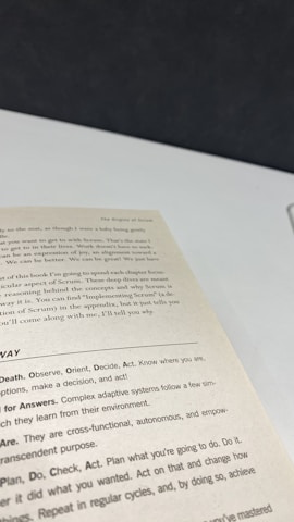 A close-up view of an open book page showing text on a desk or table. The text contains several sections with headers such as 'The Origins of Scrum' and various advice points, including observe, orient, decide, and act. The surface underneath the book is white with a dark background.