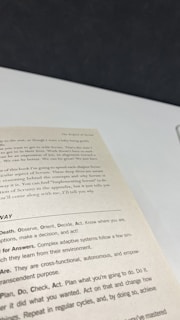 A close-up view of an open book page showing text on a desk or table. The text contains several sections with headers such as 'The Origins of Scrum' and various advice points, including observe, orient, decide, and act. The surface underneath the book is white with a dark background.