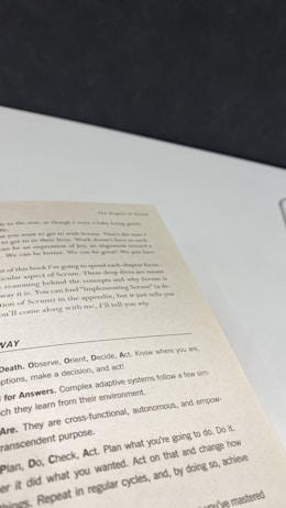 A close-up view of an open book page showing text on a desk or table. The text contains several sections with headers such as 'The Origins of Scrum' and various advice points, including observe, orient, decide, and act. The surface underneath the book is white with a dark background.