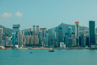 A cityscape featuring a skyline of modern skyscrapers set against a backdrop of mountains. The buildings display various corporate logos and names. The foreground includes a body of water with several small boats.