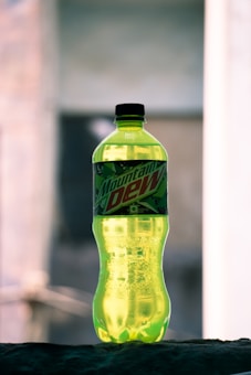 A green plastic bottle with a black cap placed on a surface. The bottle has a label with green and black patterns and the text 'Mountain Dew' in bold red letters. The background is blurred, highlighting the bottle.