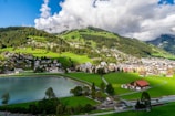 A picturesque village nestled in a lush green valley with a serene lake in the foreground. The landscape is dotted with charming houses and buildings, surrounded by rolling hills and dense forests. The sky is partly cloudy, casting dynamic shadows over the scene.