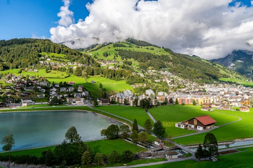 A picturesque village nestled in a lush green valley with a serene lake in the foreground. The landscape is dotted with charming houses and buildings, surrounded by rolling hills and dense forests. The sky is partly cloudy, casting dynamic shadows over the scene.