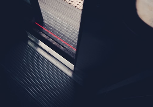 A partial view of a train or subway door with a visible threshold. The floor of the transport vehicle is metallic with grooved lines, and there is a contrasting textured platform outside. A red line runs along the edge of the platform as a warning marker.