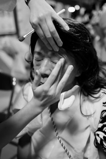A person is having makeup applied to their face by another individual's hands. The setting appears to be indoors with a slightly blurred background, possibly indicating a busy or crowded environment.