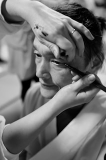 A person is having makeup applied to their eye, with a close-up focus on the hand holding an eyeliner pencil. Another hand is used to steady the face, and the environment suggests a backstage or preparation setting.