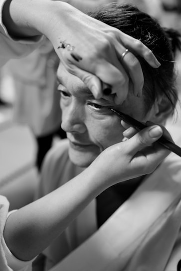 A person is having makeup applied to their eye, with a close-up focus on the hand holding an eyeliner pencil. Another hand is used to steady the face, and the environment suggests a backstage or preparation setting.