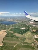 An aerial view of a vast landscape featuring patchwork fields of varying colors and shapes, a large body of water, and a distant mountain range. The airplane wing is prominently visible in the foreground, indicating the photo was taken from an in-flight perspective. The sky is clear with some scattered clouds.